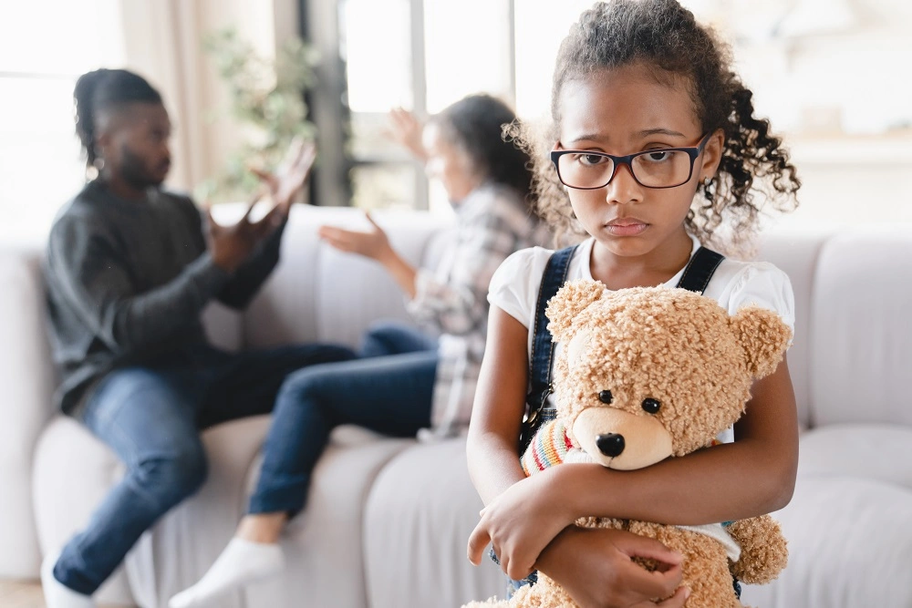 Girl holding a teddy bear while her parents argue behind her