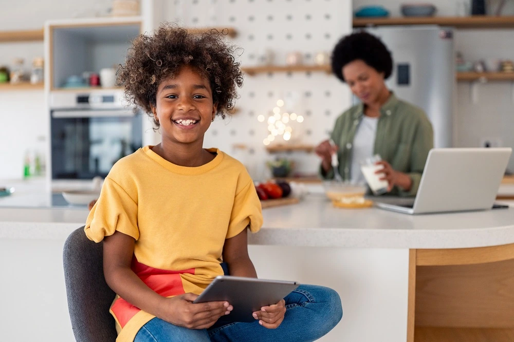 Smiling girl holding a tablet in the kitchen while her mother works behind her